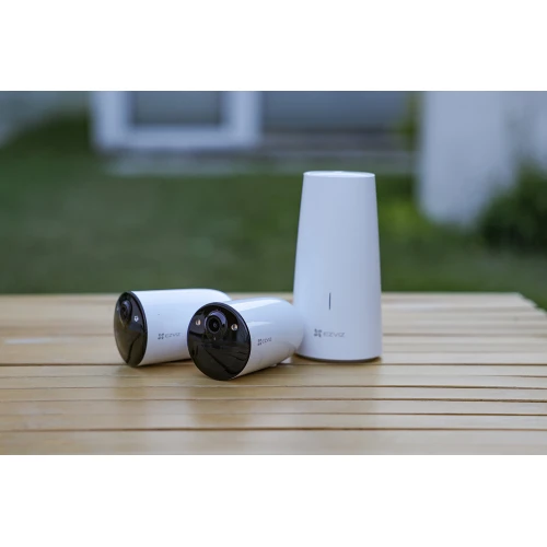 Na zdjęciu znajdują się dwie białe kamery monitoringu oraz biały, stożkowaty router, ustawione na drewnianym stole na tle rozmytego ogrodu. Kamery mają czarne, błyszczące fronty z widocznymi obiektywami.