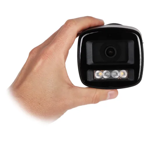 Osoba trzyma w dłoni czarną kamerę monitoringu z widocznym obiektywem i trzema diodami LED. Kamera ma prostokątny kształt i jest skierowana w stronę obiektywu aparatu.