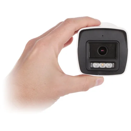 Osoba trzyma w dłoni kamerę monitoringu o kwadratowym kształcie z widocznym obiektywem i diodami LED. Kamera ma czarny front i białą obudowę.