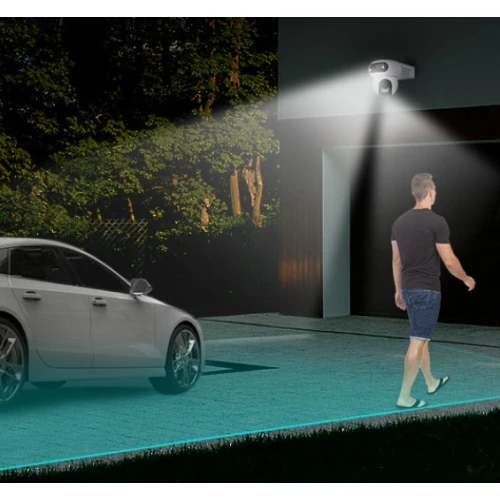 Na zdjęciu widać kamerę obrotową zamontowaną na zewnętrznej ścianie budynku, oświetlającą podjazd. W tle znajduje się zaparkowany samochód, a po prawej stronie mężczyzna idzie w kierunku budynku.