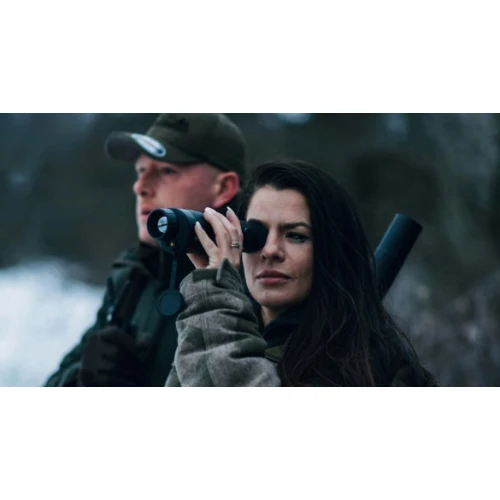 Na zdjęciu widać dwie osoby w zimowym otoczeniu, z których jedna trzyma lornetkę i patrzy przez nią, a druga ma na ramieniu broń. Obie osoby są ubrane w ciepłe, outdoorowe ubrania, a tło stanowi rozmyty, zimowy krajobraz.