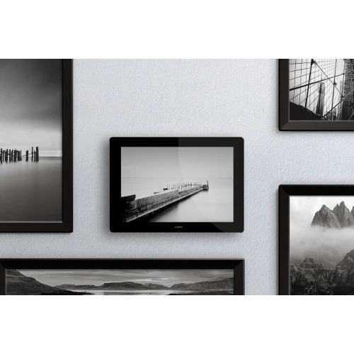 Na zdjęciu znajduje się sterownik gestem Fibaro Swipe, umieszczony na ścianie wśród czarno-białych fotografii w ramkach. Urządzenie ma elegancki, czarny design i przypomina nowoczesną ramkę na zdjęcia.