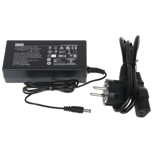 Na zdjęciu znajduje się czarny zasilacz z przewodem, zakończony wtyczką do gniazdka elektrycznego oraz wtykiem do urządzenia. Na obudowie zasilacza widoczne są różne oznaczenia i specyfikacje techniczne.