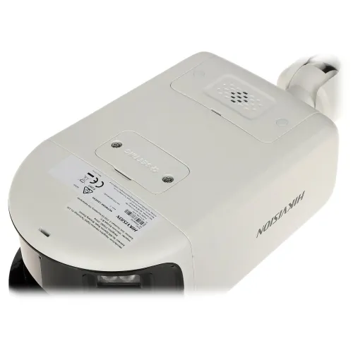 Zdjęcie przedstawia widok od spodu białego urządzenia elektronicznego marki Hikvision, z widocznymi etykietami i otworami wentylacyjnymi. W tle widać fragment kabla zasilającego.