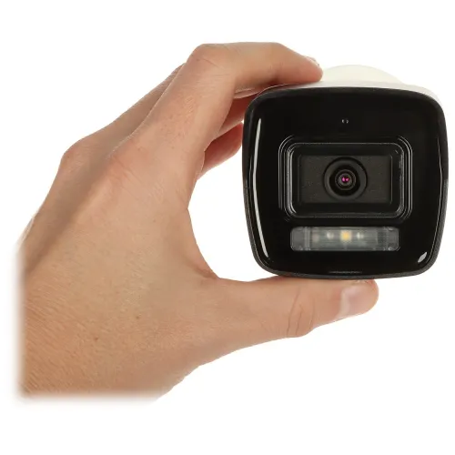 Na zdjęciu widać rękę trzymającą kamerę bezpieczeństwa, ukazaną z przodu. Kamera ma czarny front z widocznym obiektywem i diodą LED poniżej.